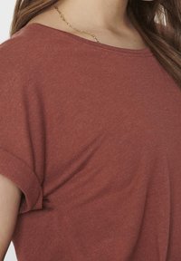 Rostfärgad löst sittande t-shirt med rullade ärmar och mjukt, texturerat tyg. Har en subtil detalj vid halslinjen med en guldkedja.