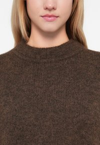 Sweater de malha castanho com gola alta e textura canelada. A imagem em primeiro plano do tecido revela padrões finos e uma aparência suave.