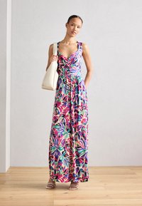 Maxi abito con un vivace e colorato motivo a foglie in verde, rosa e viola, con un corpetto stile avvolto e vita cintrata. Gli accessori includono una borsa bianca.