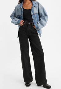 Veste en denim avec des sections bleu clair et bleu foncé, coupe oversize ; associée à un pantalon large taille haute noir et des chaussures noires.