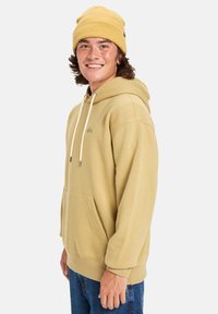 Sudadera amarilla con capucha, bolsillo frontal, cordones crema y un detalle de logo sutil. Combinada con jeans azules y un gorro a juego.