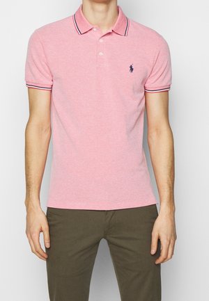 Man draagt een roze polo met korte mouwen, een marineblauw geborduurd logo en een gestreepte kraag, gecombineerd met olijfgroene broek.