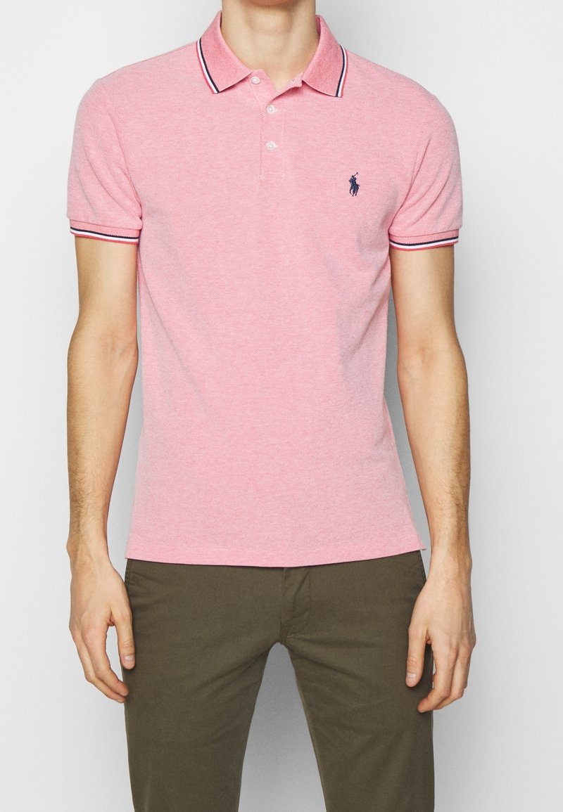 Homme portant un polo à manches courtes rose avec un logo brodé bleu marine et un col rayé, associé à un pantalon vert olive.