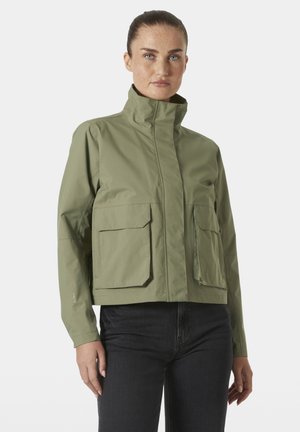 Mujer con el cabello recogido, vistiendo una chaqueta verde oliva de cuello alto con grandes bolsillos delanteros y vaqueros oscuros, de pie frente a un fondo blanco.