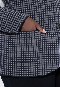 Blazer a righe navy e grigio con design a quadri, dotato di tasca frontale e chiusura a bottone. Texture morbida visibile.