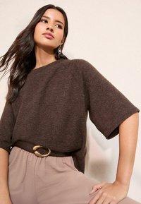 Pruun lühikeste varrukatega kootud kampsun, kombineeritud heleda beežika voltidega pükstega ja laia pruuni vöögiga, millel on kuldne pandl.