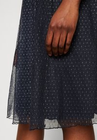 Robe bleu marine en tissu transparent texturé avec des paillettes argentées, longueur genoux, et bordure en forme de vagues. Une main repose sur le tissu.
