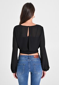 Schwarzes Chiffon-Crop-Top mit langen Ärmeln, Details mit Schlüsselverschluss am Rücken, kombiniert mit blauen Denim-Jeans mit fünf Taschen und einem braunen Leder-Patch.