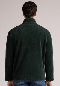 Ciemnozielony polar z wysokim kołnierzem, o miękkiej fakturze i długich rękawach. Posiada luźny krój i swobodną sylwetkę.