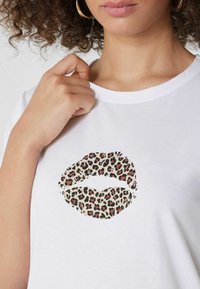 Camiseta blanca con un gráfico de beso de estampado de leopardo. Tejido suave, cuello redondo y un corte relajado. Mano descansando cerca del cuello.