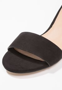 Vorderansicht Nahaufnahme einer schwarzen Wildleder-Sandalette mit offenem Zehenbereich und einem breiten Riemen über den Zehen sowie einer beigen Innensohle.