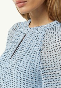 Lichtblauwe blouse met een geometrisch bladdessin in donkerblauw. Het ontwerp heeft een ronde halslijn en subtiele plooidetails.