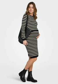 Robe de maternité rayée noire et beige, cintrée avec manches longues, associée à des bottines noires et un sac à main noir.