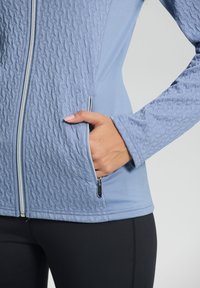 Helles blaues Zip-Hoodie mit strukturiertem Muster auf der Vorderseite und an den Ärmeln, ausgestattet mit Seitentaschen und einer glatten Stoffoberfläche.