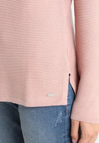 Light pink ribbed knit sweater with long sleeves, side slits, and a small silver logo tag. Paired with denim jeans.
