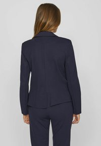 Marineblå skræddersyet blazer med lange ærmer og en struktureret pasform. Har en klippet revers, en glat tekstur og sømdetaljer langs ryggen.