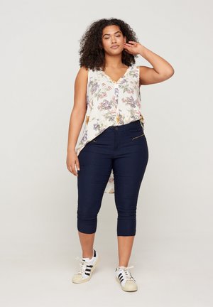 Weißes Blumenshirt mit Knopfleiste, kombiniert mit dunkelblauen, knöchelfreien Hosen und weißen Sneakers mit schwarzen Streifen.