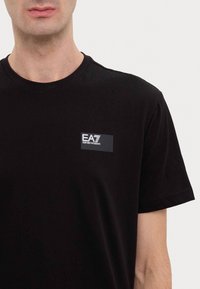 Juodas medvilnės marškinėliai su mažu stačiakampiu logotipu viršuje kairėje, kuriame pilka tekstas ir balti akcentai. Trumpomis rankovėmis ir apvaliu kaklu.