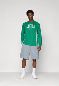 Green long-sleeve Nike shirt with "CELTICS" in white, paired with gray athletic shorts. Model wears white sneakers and crew-length socks.