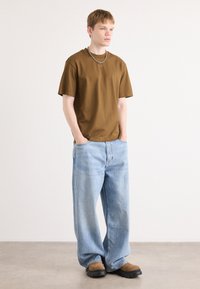 Braunfarbiges Baumwoll-T-Shirt, locker geschnitten, kombiniert mit hellblauen, weit geschnittenen Denim-Jeans. Schwarze und braune klobige Schuhe runden den Look ab.