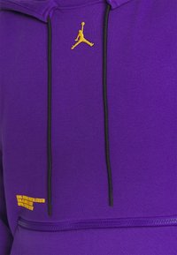 Sudadera morada con cordón negro, bordado del logo de Jordan en amarillo y texto impreso en amarillo en la parte inferior frontal. Textura de tela suave.