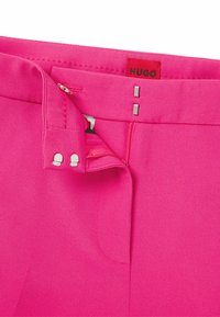 Fuksia-housut, jotka on valmistettu sileästä kankaasta ja joissa on piilovetoketju, koukku kiinnitys sekä hopeiset yksityiskohdat. Housuissa on etiketti, jossa lukee "HUGO."