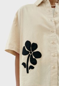 Short-sleeved beige button-up shirt featuring a large black floral embroidery design on the left side. Soft fabric with a smooth texture.