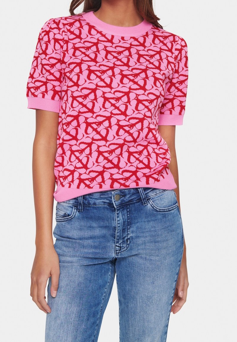 Pink knitted sweater featuring a red abstract pattern, short sleeves, and a round neck, paired with light blue denim jeans.