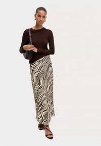 Haut marron à manches longues associé à une jupe longue beige fluide ornée de motifs ondulés noirs, accessoirisé d'un petit sac noir et de sandales à lanières.