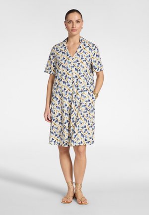 Vrouw die een knielang, kort mouw jurkje draagt met blauwe en gele bladpatronen, gouden sandalen, gouden oorbellen en een gouden armband.