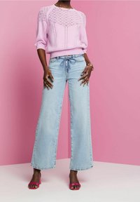 Pull en maille rose clair avec détails texturés, associé à un jean large bleu clair. Le mannequin porte des sandales à talons roses. Le fond est rose.