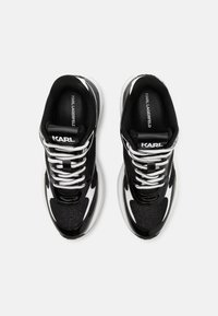 Chaussures de sport noires avec des accents blancs, tige en mesh, semelle épaisse et design volumineux. Présente des lacets noirs et une étiquette logo sur la languette.
