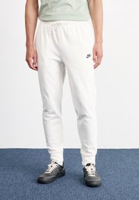 Białe dresy Nike z elastycznym paskiem w talii i sznurkiem do regulacji. Zawierają małe czarne logo. Model nosi czarne sneakersy na niebieskim tle.