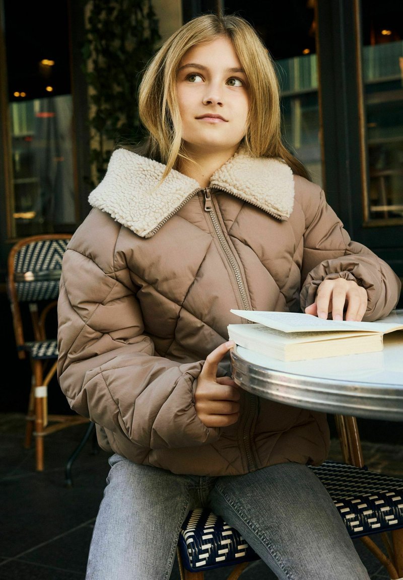 Jeune personne portant une veste matelassée marron assise à une table de café en train de lire un livre, regardant légèrement sur le côté avec une expression pensive.