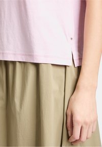 T-shirt en coton rose avec fente sur le côté, agrémenté d'un petit bouton décoratif près de l'ourlet. Associé à une jupe plissée beige fluide.