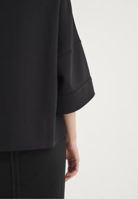 Černá blůza se širokými rukávy, vyrobená z hladkého materiálu, s zaobleným spodním lemem. Dolní okraj má jemný šev. Ruka držící černou sukni.