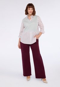 Camicia a righe bianca e rossa con un gilet di maglia grigia sovrapposto, abbinata a pantaloni larghi bordeaux e tacchi nudi. Tessuti lisci e design casual.