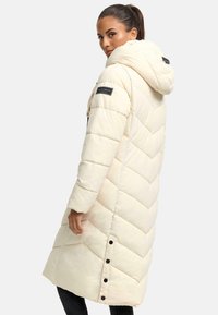Long, puffy cream-colored coat with a hood, chevron pattern, and black buttons along the side. Features a branded patch on the sleeve.