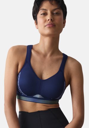 Kvinde med kort mørkt hår iført en tætsiddende marineblå sports-bh og sorte højtaljede leggings mod en ensfarvet lys baggrund.
