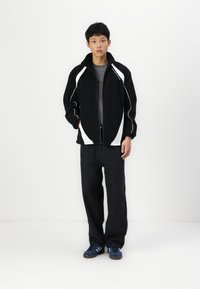 Giacca nera con zip e accenti bianchi, abbinata a pantaloni scuri e sneakers blu. La giacca presenta un colletto alto e una vestibilità comoda.