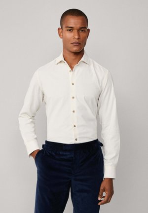 Camisa blanca de manga larga con cuello, adornada con botones marrones, combinada con pantalones de pana azul marino. Textura de tela suave.