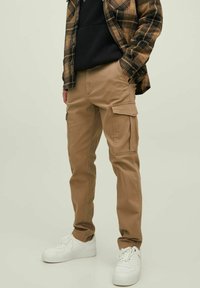 Persona che indossa pantaloni cargo color beige, sneakers bianche, felpa nera e giacca a quadri marrone con una mano in tasca, su uno sfondo semplice.