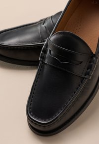 Paire de mocassins noirs en cuir sans lacets avec des détails cousus et une bride décorative sur le dessus, placés sur une surface beige.