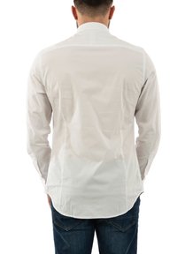 Camisa blanca de manga larga con cuello puntiagudo, que presenta una textura suave y un dobladillo ligeramente curvado, llevada sobre unos pantalones vaqueros de mezclilla azul.