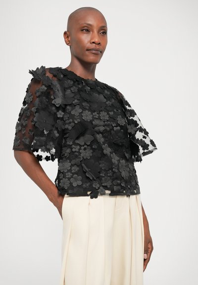 Sort bluse lavet af gennemsigtigt stof, prydet med teksturerede blomsterapplikationer. Har korte ærmer og rund halsudskæring. Parret med cremefarvede bukser.