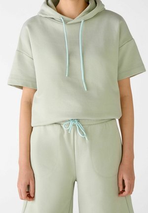 Henkilö, joka pukeutunut vaaleanvihreään lyhythihaiseen huppariin ja vastaaviin nyörisäärystimiin, kädet rentoina sivuilla, valkoisella taustalla.