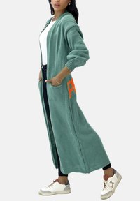 Lång, grön cardigan i stickad tyg med öppen framsida, två fickor och orange textdetalj. Matcher med svarta jeans och vita sneakers.