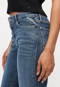 Κοντινό πλάνο ενός ατόμου που φοράει μπλε τζιν Replay και μαύρο cropped πουλόβερ, τονίζοντας την υφή του τζιν και τις λεπτομέρειες στις τσέπες.