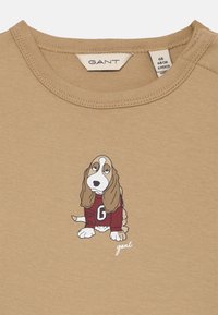 Lysbrun sweatshirt med et basset hound-tryk, der bærer en maroon sweater med et 'G' på. Etiketten viser "GANT" med størrelsesoplysninger.