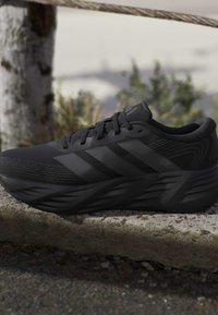 Black athletic running shoe with three stripes on the side, resting on a concrete ledge outdoors with blurred greenery in the background.
